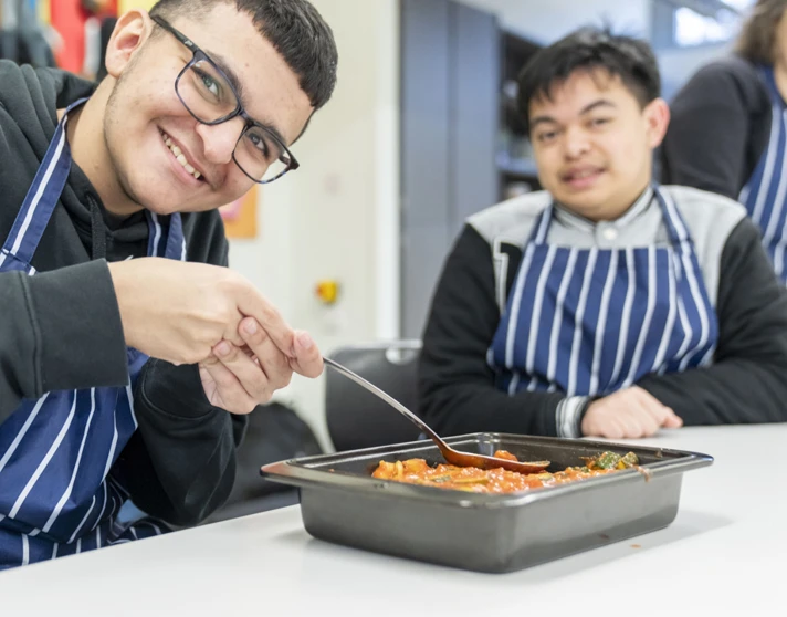 Two young men smiling and preparing a tray of food, wearing striped aprons in a kitchen setting. Two young men smiling and preparing a tray of food, wearing striped aprons in a kitchen setting.